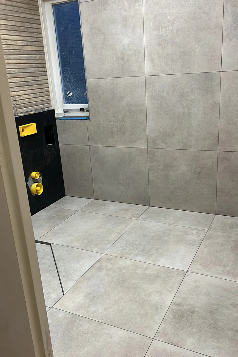 Strak tegelwerk met 90x90 cm tegels op vloer en wand in badkamer met toilet, uitgevoerd door Roy van den Bosch – VDB Tegelwerk.