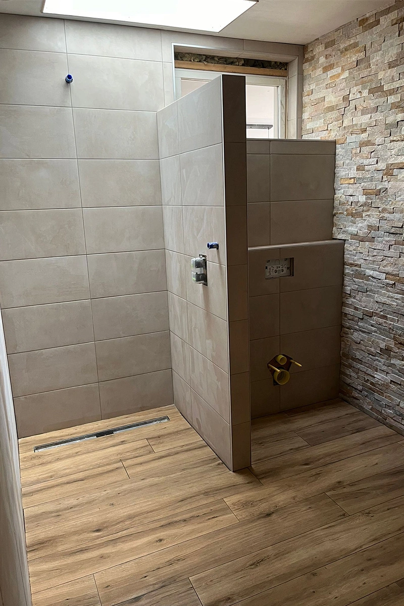 Badkamer en toilet met houtlook vloertegels, strakke neutrale wandafwerking, schuine vloer met afschot voor douche en natuursteen mozaïek accenten. Lichte ruimte dankzij lichtkoepel in het plafond. Tegelwerk door VDB Tegelwerk.