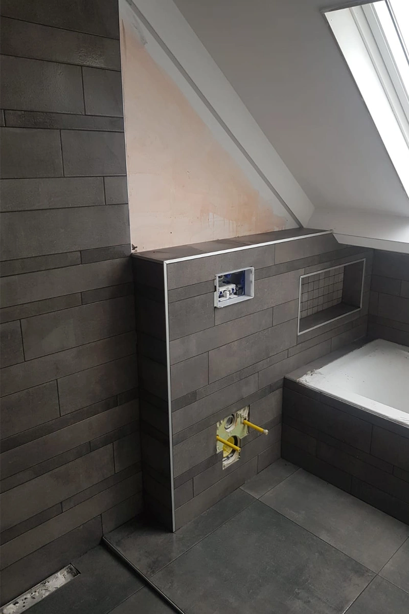 Badkamer met donkere tegels, schuin afschot bij de douche, een ligbad, ingebouwde nisjes en een speels effect door gebruik van verschillende tegelformaten. Vakwerk door VDB Tegelwerk.