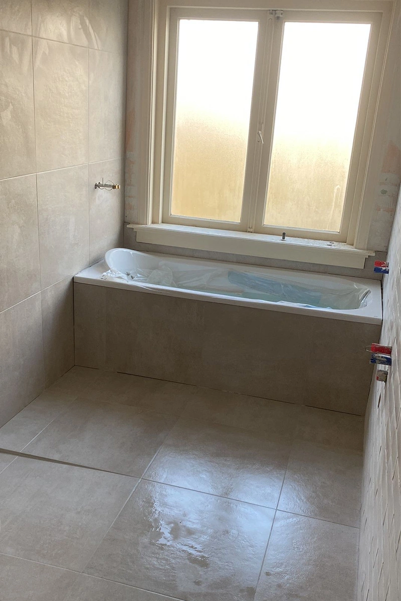 Lichte badkamer met ligbad, ruime douchezone met schuin afschot en zachte, neutrale tegelkleuren. Afwerking door VDB Tegelwerk.