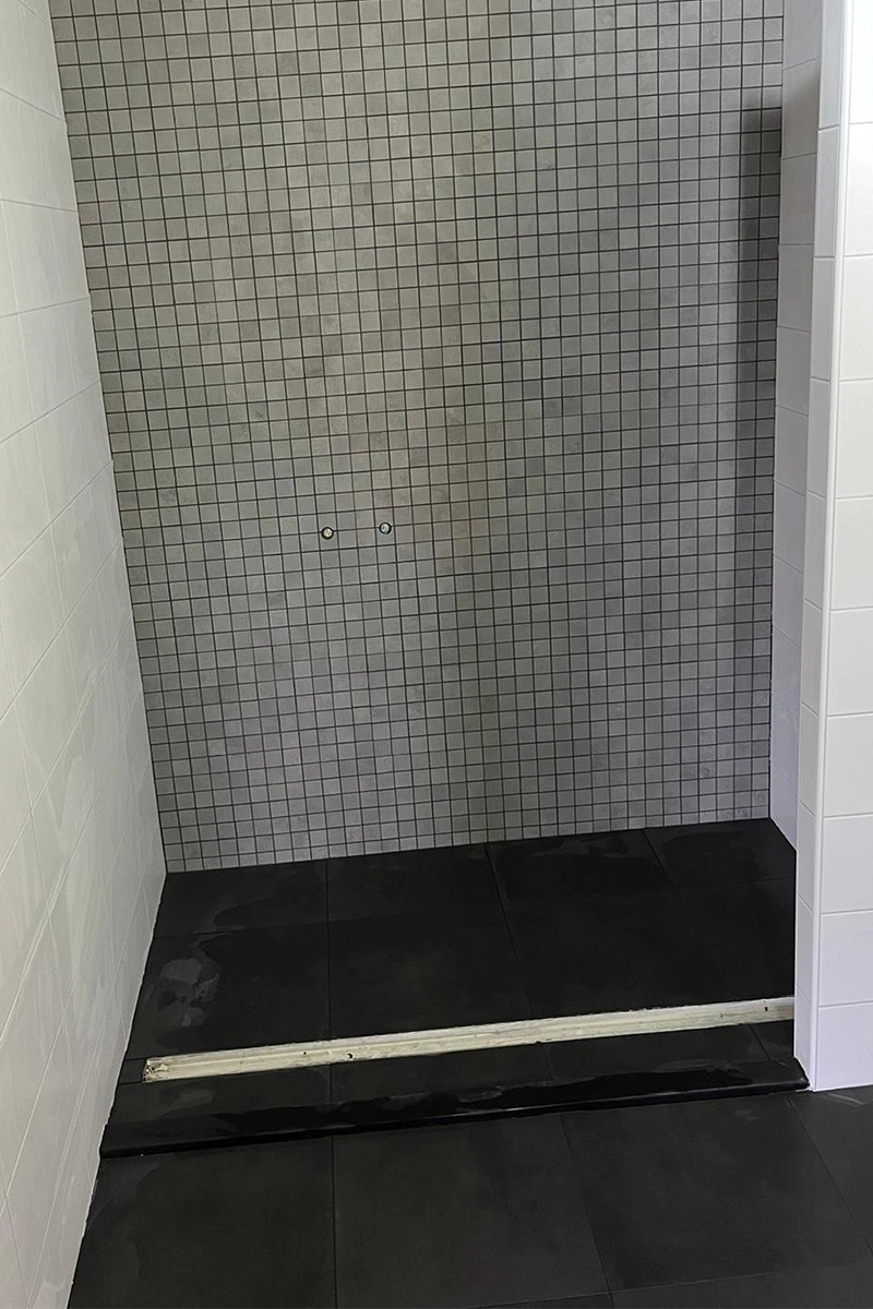 Ruime moderne douchehoek met robuuste zwarte vloertegels, kleine grijze mozaïektegels aan de muur en een strak wit douchewandje met witte wandafwerking. Uitgevoerd door VDB Tegelwerk.